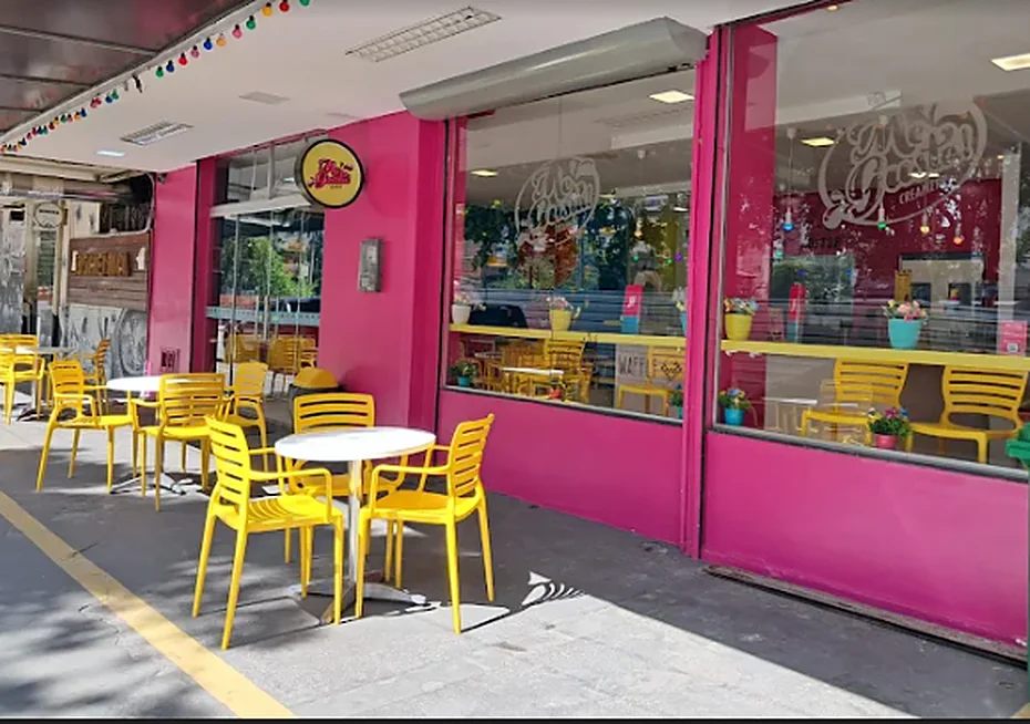 Fachada rosa da Me Gusta com mesas e cadeiras amarelas na calçada